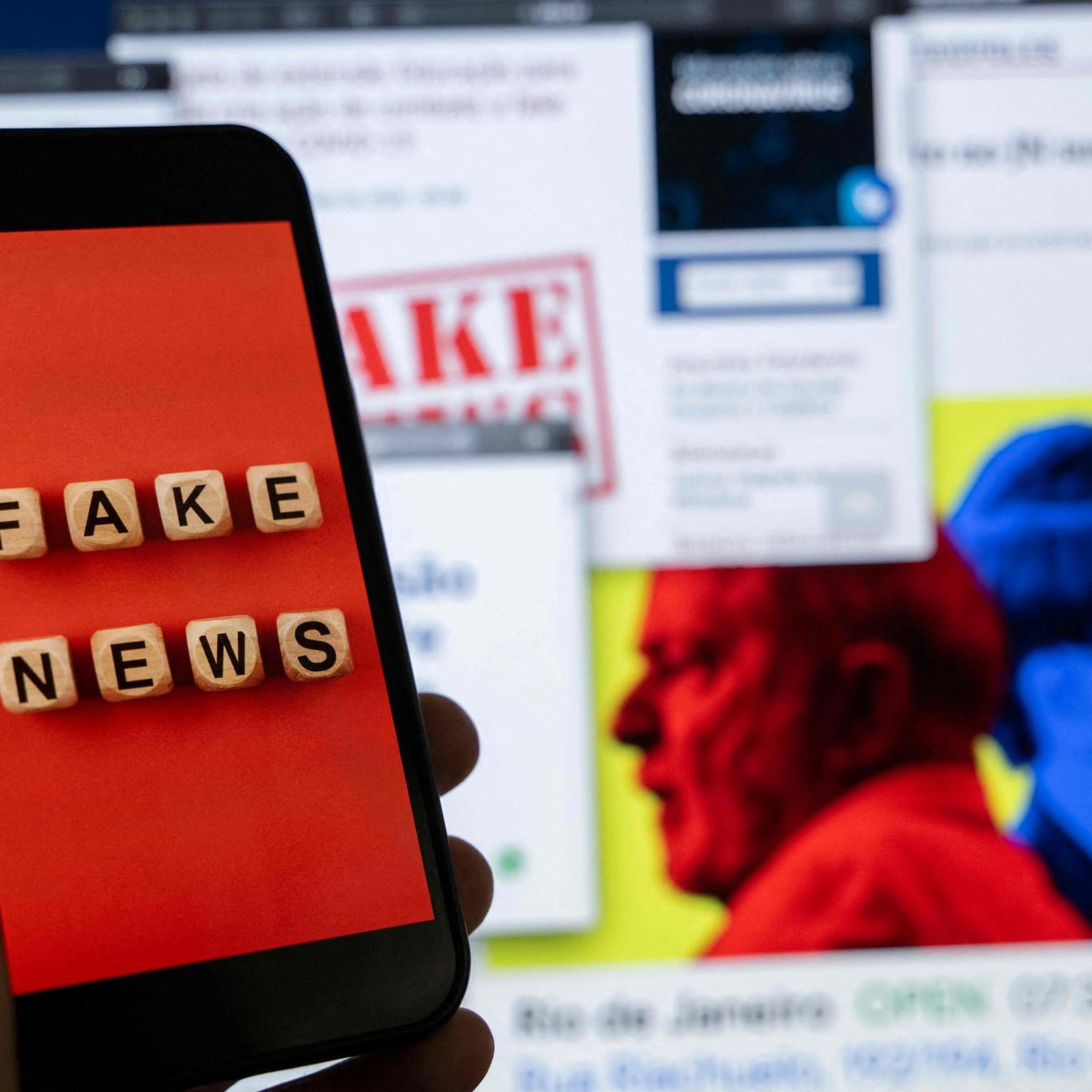 Vor einem Computerbildschirm wird ein Smartphone hochgehalten, auf dem "Fake News" zu lesen ist