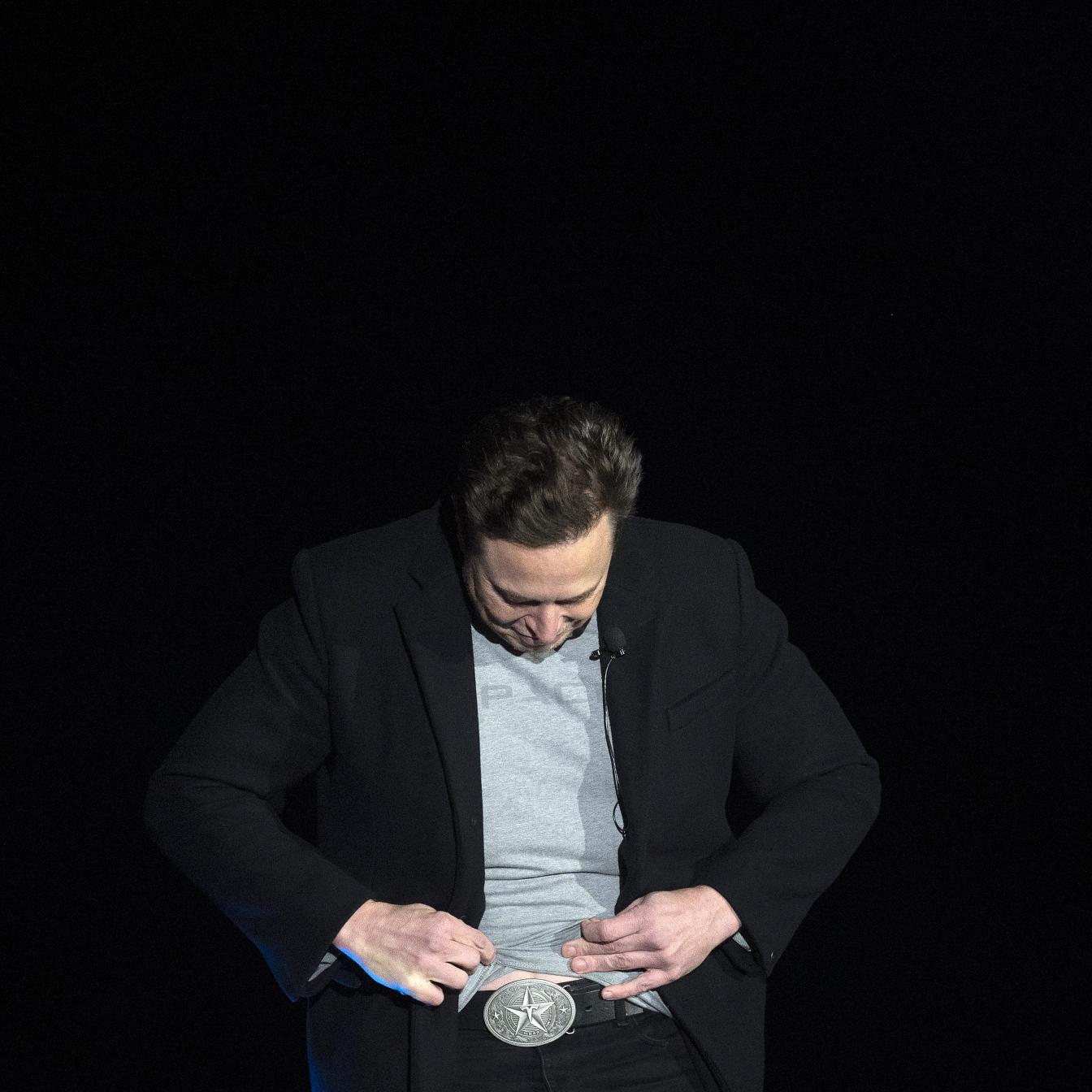 Elon Musk schaut während einer Pressekonferenz auf seine Gürtelschnalle